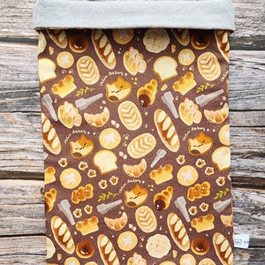 Peut inclure: Sac en tissu marron avec un motif répété de divers produits de boulangerie, notamment du pain, des croissants et des pâtisseries. Le sac a un dessus plié gris clair. Le texte "calico bakery" est imprimé.