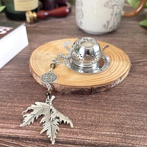 May include: A silver-toned metal tea infuser shaped like a miniature teapot, resting on a small circular tray. A decorative chain is attached, featuring a round filigree charm, two dark brown beads, and a large silver-toned maple leaf pendant.