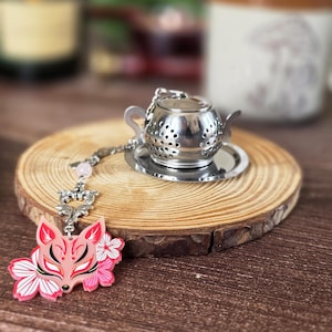Puede incluir: Un infusor de té de metal plateado con forma de tetera en miniatura sobre un platillo pequeño. Una cadena plateada está unida al asa, con una cuenta rosa claro y un dije plano de máscara de zorro rosa decorado con patrones florales.