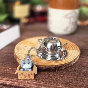 Può includere: Un infusore per tè in metallo argentato a forma di teiera in miniatura, poggiato su un piattino circolare abbinato. Una catenella argentata collega l'infusore a una piccola statuetta di gatto grigio seduto in una scatola di cartone beige con testo rosso.