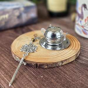 Pode incluir: Um infusor de chá de metal prateado em forma de bule em miniatura sobre um pires a condizer, acompanhado por uma corrente decorativa com um pendente de flor em filigrana e uma espada longa e fina com acabamento metálico.