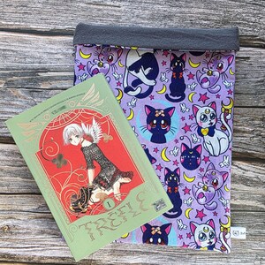 May include: A green manga book with red detailing and gold lettering reading "TRC" sits beside a purple book sleeve. The sleeve has a pattern of cartoon cats, moons, and stars. The top of the sleeve is grey.