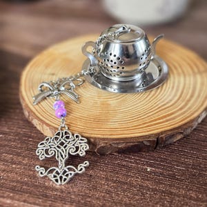 Può includere: Un infusore per tè in metallo argentato a forma di teiera in miniatura, poggiato su un piattino circolare abbinato. Una catenella argentata fissata al manico presenta un fiocco decorativo in metallo, due piccole perline viola e un ciondolo.