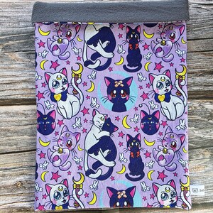 May include: A lavender fabric sleeve with a repeating pattern of cartoon cats, moons, and stars. The cats are in various poses, some with bows and crescent moon symbols. The top edge is a solid grey fabric.