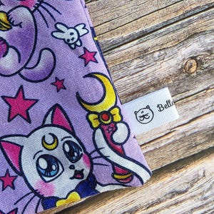 May include: A close-up of a purple fabric with a repeating pattern of cartoon cats, stars, and crescent moons. The fabric also features a white bunny and a wand. A small white tag with the word "Belle" and a cat logo is visible.