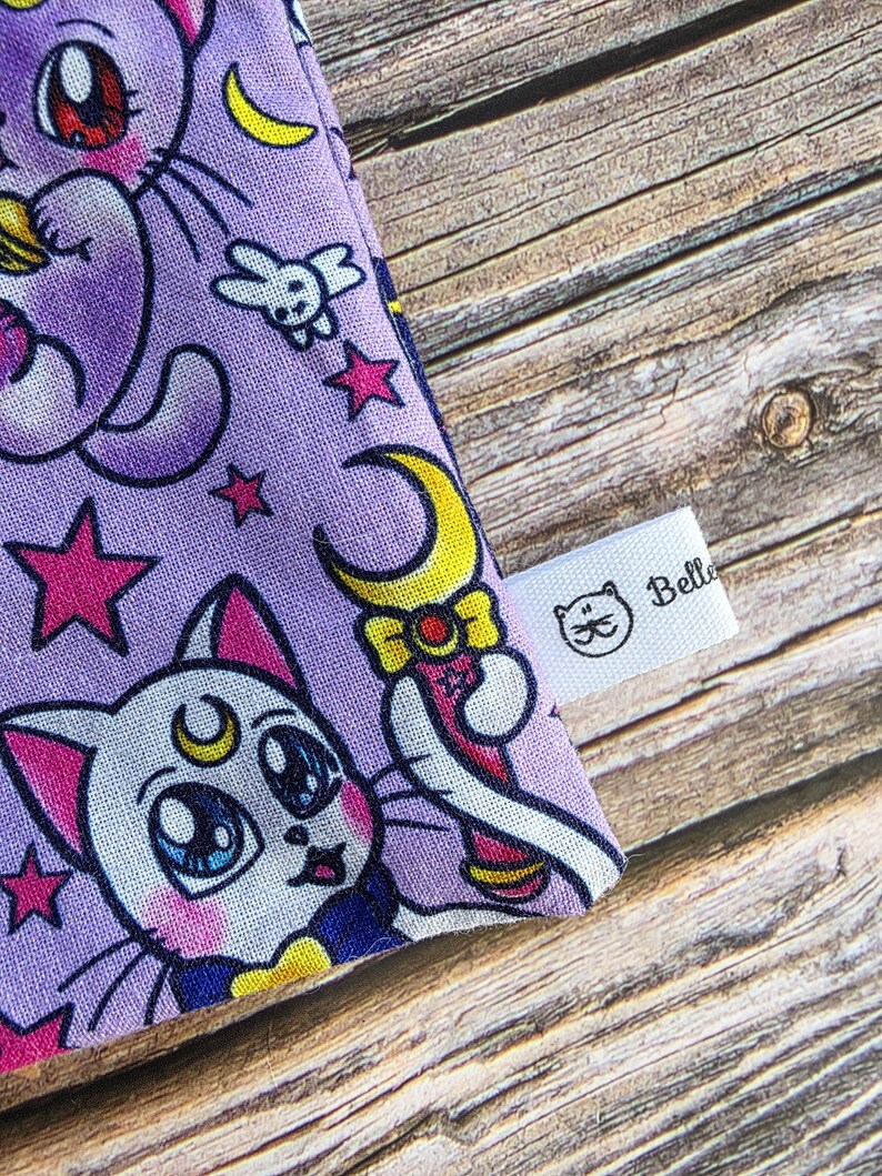 May include: A close-up of a purple fabric with a repeating pattern of cartoon cats, stars, and crescent moons. The fabric also features a white bunny and a wand. A small white tag with the word "Belle" and a cat logo is visible.