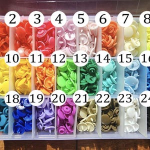 May include: A clear plastic organiser filled with colourful snap fasteners. The snaps are arranged in rows by colour, including red, orange, yellow, green, blue, and purple. Each compartment is labelled with a number.