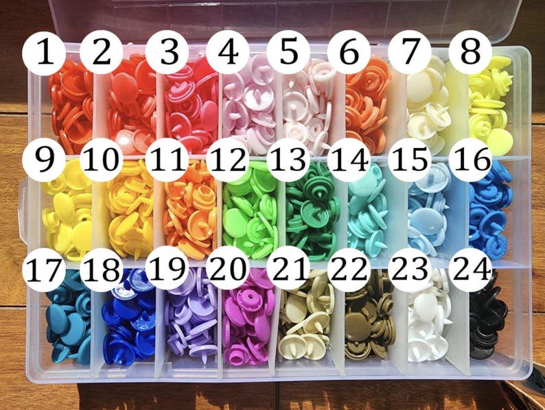 May include: A clear plastic organiser filled with colourful snap fasteners. The snaps are arranged in rows by colour, including red, orange, yellow, green, blue, and purple. Each compartment is labelled with a number.