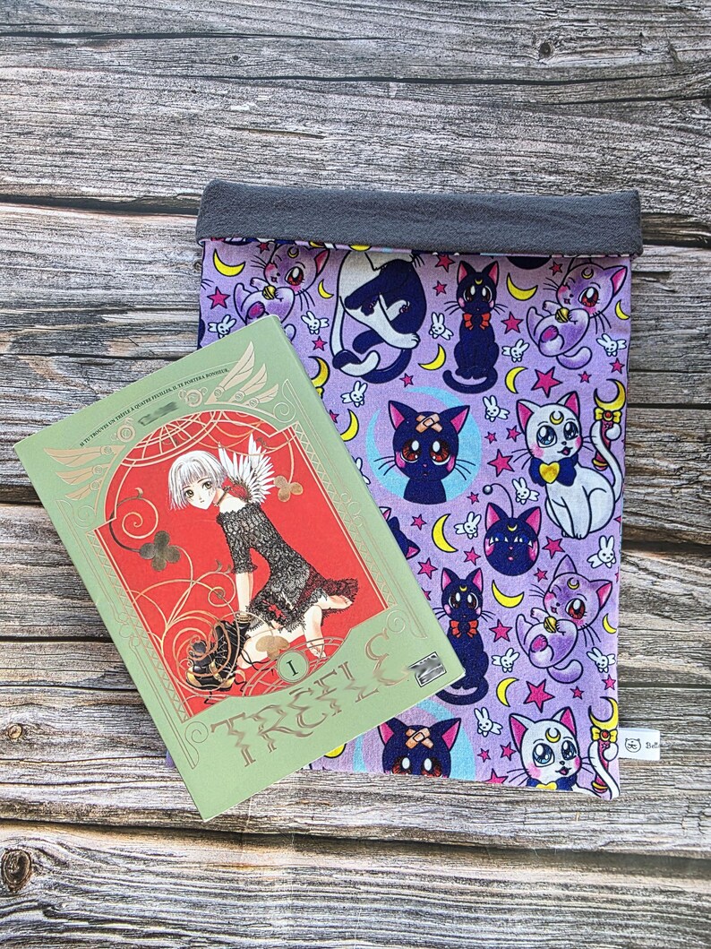May include: A green manga book with red detailing and gold lettering reading "TRC" sits beside a purple book sleeve. The sleeve has a pattern of cartoon cats, moons, and stars. The top of the sleeve is grey.