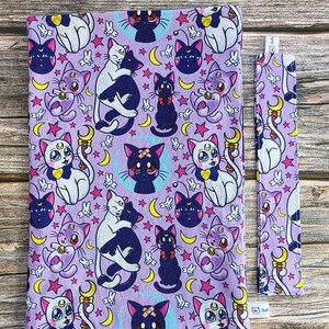 May include: A lavender book cover and bookmark with a repeating pattern of cartoon cats, moons, stars, and other celestial elements. The cats are in various poses, with white, black, and purple colouring. The bookmark is a long, narrow rectangle.