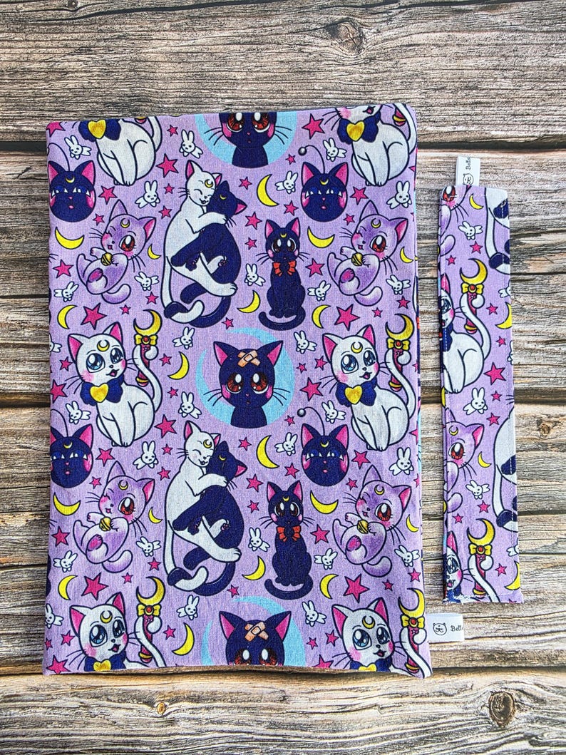 May include: A lavender book cover and bookmark with a repeating pattern of cartoon cats, moons, stars, and other celestial elements. The cats are in various poses, with white, black, and purple colouring. The bookmark is a long, narrow rectangle.