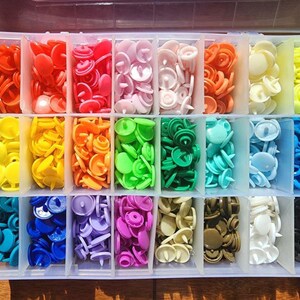 May include: A clear plastic storage box containing a rainbow of snap fasteners. The colours include red, pink, orange, yellow, green, blue, purple, brown, white, and black. Each colour is stored in a separate compartment.