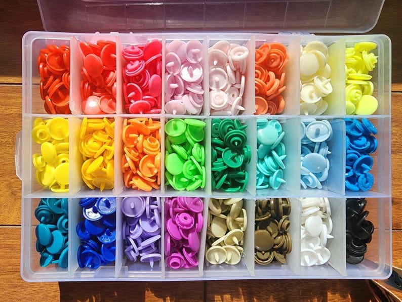 May include: A clear plastic storage box containing a rainbow of snap fasteners. The colours include red, pink, orange, yellow, green, blue, purple, brown, white, and black. Each colour is stored in a separate compartment.