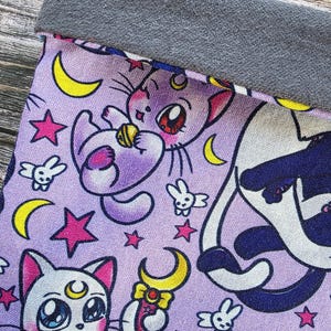 May include: A lavender fabric featuring a repeating pattern of cartoon cats, crescent moons, stars, and small bunny figures. The top edge is a solid grey fabric. The cat illustrations have large eyes and are in various poses.
