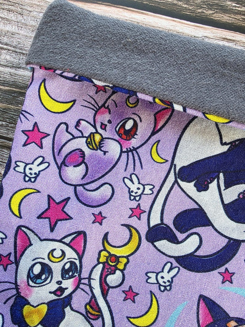 May include: A lavender fabric featuring a repeating pattern of cartoon cats, crescent moons, stars, and small bunny figures. The top edge is a solid grey fabric. The cat illustrations have large eyes and are in various poses.