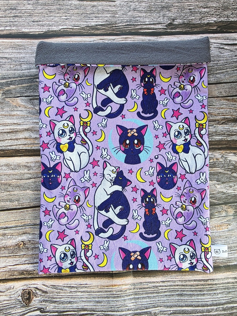 May include: A lavender fabric sleeve with a repeating pattern of cartoon cats, moons, and stars. The cats are in various poses, some with bows and crescent moon symbols. The top edge is a solid grey fabric.