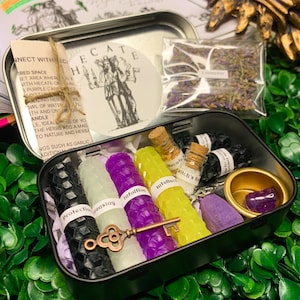 May include: A metal tin filled with various witchy items, including candles, herbs, and a small vial. The tin is labeled "Hecate" and has a drawing of a woman with a crown and a staff. The candles are labeled "protection", "cleansing", "intuition", and "intellect". The herbs are labeled "lavender" and "witch's salt".