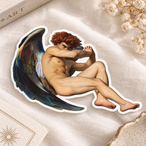 May include: A die-cut sticker featuring a figure with auburn hair and large, dark blue and iridescent wings. The figure is depicted in a curled, seated position with arms crossed, showcasing detailed musculature and light skin tones.