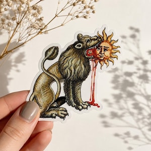 May include: A die-cut sticker featuring an alchemical illustration of a brown lion with a dark mane biting into a golden sun with a human face, with red liquid dripping from the sun, rendered in a detailed, vintage etching art style.
