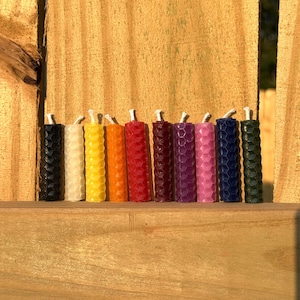 May include: A collection of ten colorful, honeycomb-patterned candles. The candles are arranged in a row, showcasing a spectrum of colors including black, white, yellow, orange, red, burgundy, purple, pink, navy blue, and green. Each candle has a white wick.
