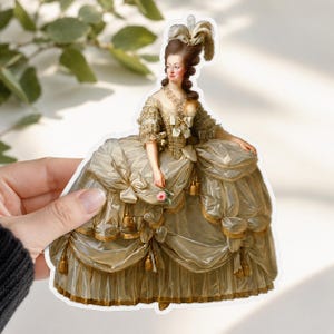 Peut inclure: Un autocollant découpé représentant un portrait d'une personne en robe de style XVIIIe siècle. La silhouette porte une coiffure haute poudrée avec une plume et une robe volumineuse aux reflets dorés, ornée de pompons et d'une petite rose rose.