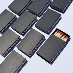May include: An overhead shot of several closed and one open black matchboxes. The open box reveals red-tipped matches. The matchboxes are arranged on a light blue surface. The striking strip is visible on the side of the boxes.