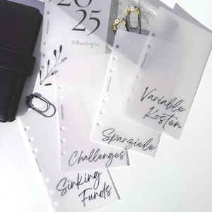 Könnte beinhalten: Transparente Planer-Einlagen mit Beschriftungen wie "Sinking Funds" und "Challenges". Die oberste Einlage zeigt "2025" und "Two thousand twenty-five". Ein schwarzer Lederplaner und dekorative Büroklammern sind ebenfalls zu sehen.