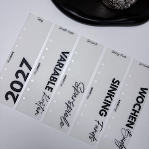Könnte beinhalten: Fünf halbtransparente, rechteckige Planer-Register aus Kunststoff mit sechs Stanzlöchern an der linken Seite. Jeder Registerstreifen ist mit schwarzem Text bedruckt, darunter 2027, Variable Kosten, Sparspiele, Sinking Funds und Wochen Budget.