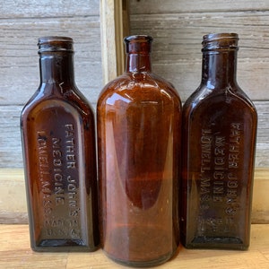 Könnte beinhalten: Drei bernsteinfarbene Glasflaschen mit rechteckiger Form und abgerundetem Boden. Jede Flasche hat ein Etikett mit dem Text "Father John's Medicine Lowell, Mass. Made in U.S.A."
