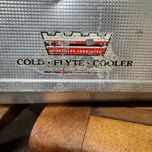 May include: Close-up of a vintage silver metal cooler with a textured surface. The cooler has a faded emblem with the words "FIBER-GLAS-INSULATED" and "COLD FLYTE COOLER." The text "Hamilton-Ketch Corp. Hamilton, Ohio" is also visible.