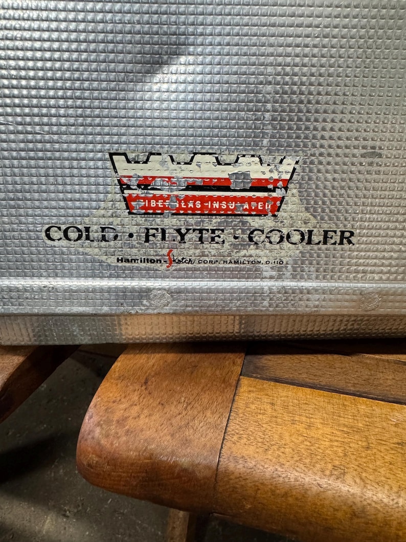 May include: Close-up of a vintage silver metal cooler with a textured surface. The cooler has a faded emblem with the words "FIBER-GLAS-INSULATED" and "COLD FLYTE COOLER." The text "Hamilton-Ketch Corp. Hamilton, Ohio" is also visible.