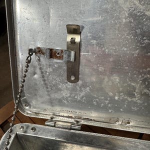 May include: An open, weathered metal container with a hinged lid. A metal bottle opener is attached to the inside of the lid. A small chain is connected to the lid. The container has a silver finish and shows signs of age and use.