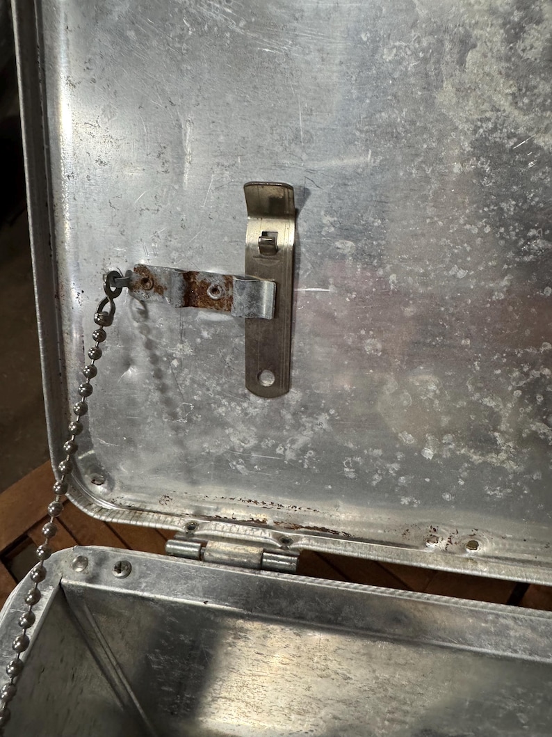 May include: An open, weathered metal container with a hinged lid. A metal bottle opener is attached to the inside of the lid. A small chain is connected to the lid. The container has a silver finish and shows signs of age and use.