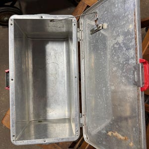 May include: Open, vintage aluminum cooler with a red handle and latch. The interior is empty and shows signs of wear. The lid is attached by a chain. The cooler is sitting on a wooden surface.
