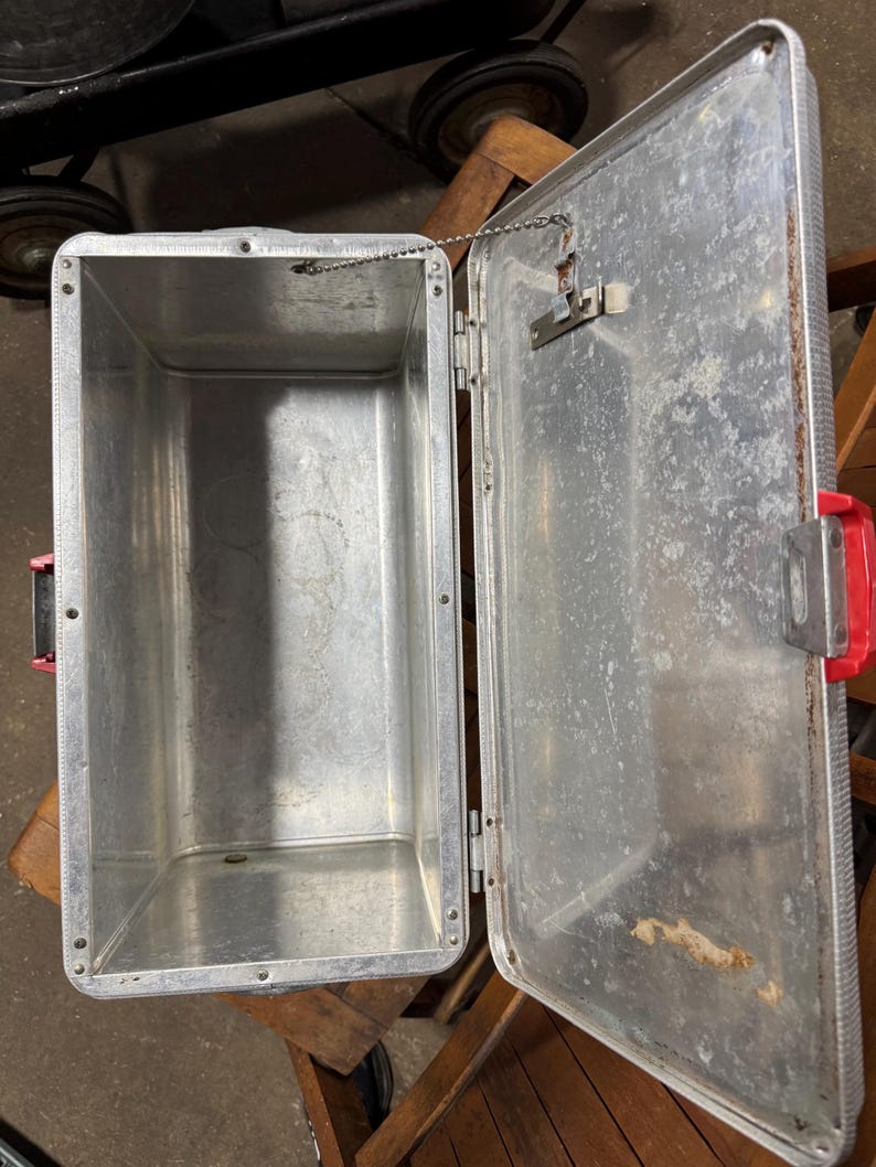 May include: Open, vintage aluminum cooler with a red handle and latch. The interior is empty and shows signs of wear. The lid is attached by a chain. The cooler is sitting on a wooden surface.
