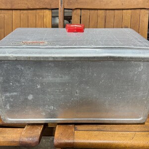May include: A vintage, rectangular metal cooler with a textured surface and a red plastic handle. The cooler is silver and shows signs of wear, suggesting age. It sits on a wooden bench.