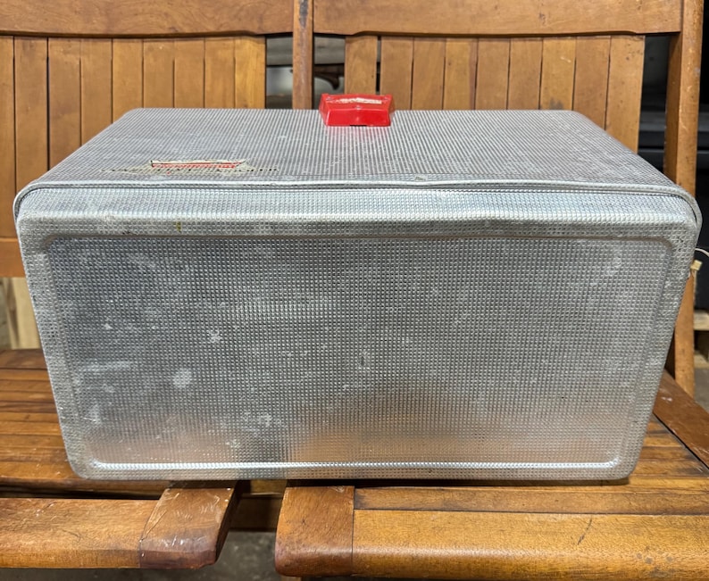 May include: A vintage, rectangular metal cooler with a textured surface and a red plastic handle. The cooler is silver and shows signs of wear, suggesting age. It sits on a wooden bench.