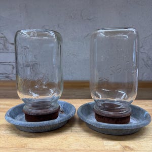 Pode incluir: Dois frascos de vidro transparente com texto em relevo, cada um sobre uma pequena bandeja redonda de metal galvanizado. Os frascos têm uma base circular castanha. O fundo é uma parede cinza claro e uma superfície de madeira.
