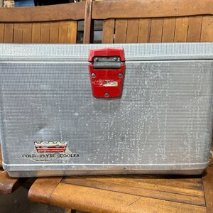 May include: A vintage silver metal cooler with a red latch. The cooler has a textured surface and a logo that reads "COLD-FLYTE COOLER". The cooler is rectangular and appears to be in good condition.