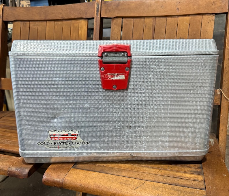 May include: A vintage silver metal cooler with a red latch. The cooler has a textured surface and a logo that reads "COLD-FLYTE COOLER". The cooler is rectangular and appears to be in good condition.