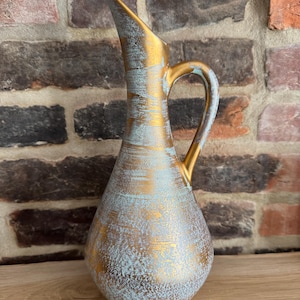 May include: A decorative gold-colored pitcher with a handle and a long, pointed spout. The pitcher has a textured surface with a turquoise and gold finish. The pitcher is a vintage-style home decor item.
