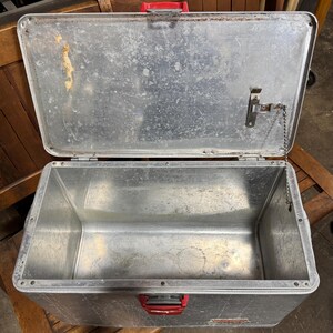 May include: An open, vintage metal lunchbox with a silver finish and red handle and latches. The lunchbox is rectangular with rounded corners and shows signs of wear and age. The lid is attached by a chain.