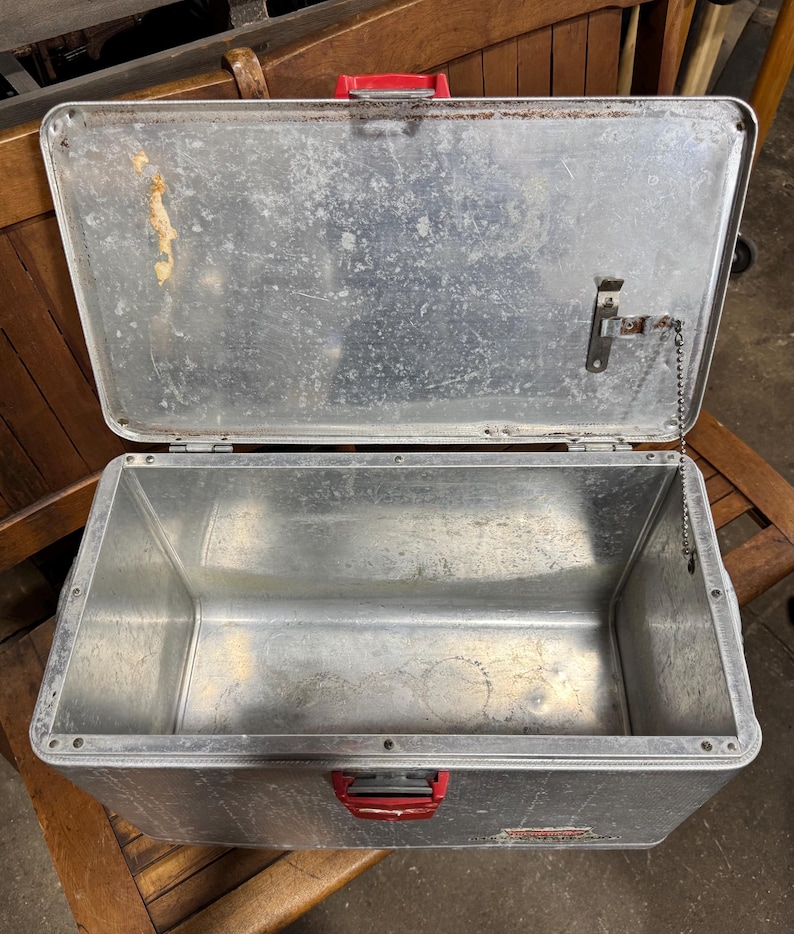 May include: An open, vintage metal lunchbox with a silver finish and red handle and latches. The lunchbox is rectangular with rounded corners and shows signs of wear and age. The lid is attached by a chain.