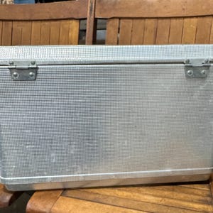 May include: A rectangular, silver-colored metal cooler with a textured surface and a hinged lid. The cooler has two metal clasps on the front and is sitting on a wooden chair. The cooler is designed to keep items cold.