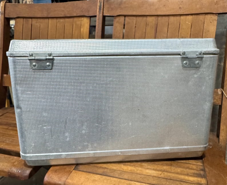 May include: A rectangular, silver-colored metal cooler with a textured surface and a hinged lid. The cooler has two metal clasps on the front and is sitting on a wooden chair. The cooler is designed to keep items cold.