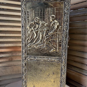 Puede incluir: Una pantalla de chimenea de latón vintage con una escena en relieve detallada de dos figuras en una habitación. La pantalla tiene un borde decorativo y un panel inferior texturizado. El latón tiene una pátina cálida y envejecida, lo que le da encanto.