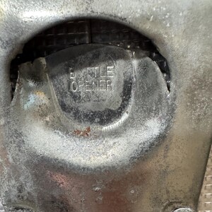 May include: Close-up of a weathered, silver-toned bottle opener. The word "BOTTLE OPENER" is embossed in the center. The opener has a rounded shape with two rivets at the bottom.