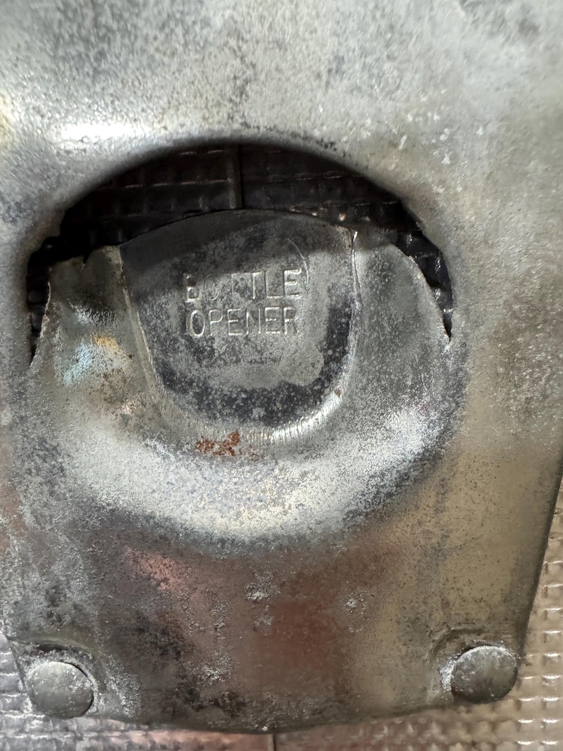 May include: Close-up of a weathered, silver-toned bottle opener. The word "BOTTLE OPENER" is embossed in the center. The opener has a rounded shape with two rivets at the bottom.
