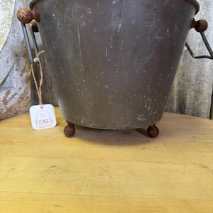 Puede incluir: Un cubo de cobre vintage con asas y patas de madera. El cubo tiene una pátina desgastada y envejecida y es perfecto para una decoración rústica o de granja.