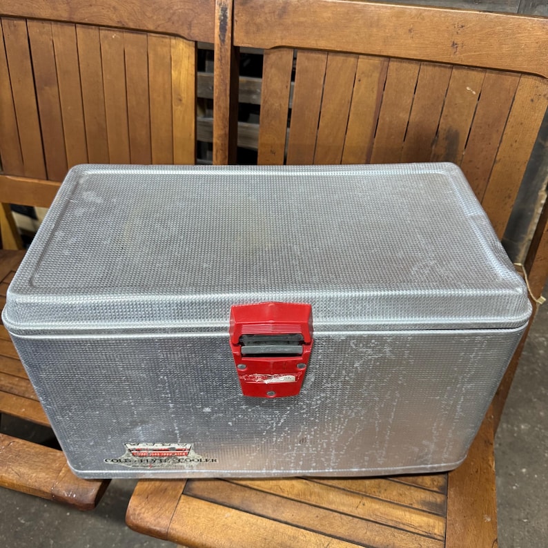 May include: A vintage silver metal cooler with a red latch. The cooler has a textured surface and a rectangular shape. The words "COLD FLYING COOLER" are printed on the front.
